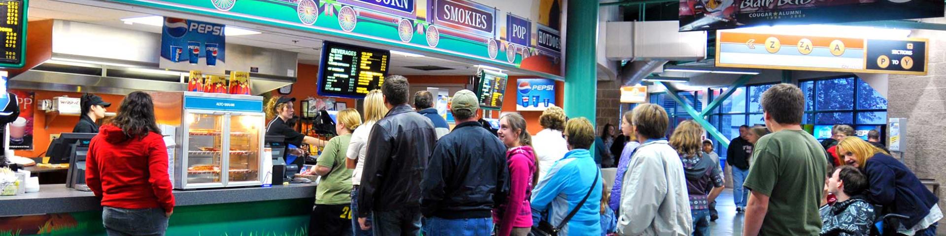 About 10 people in a lineup to purchase food at a concession. Concession features photos of foods available, including burgers, fries, and coffee.